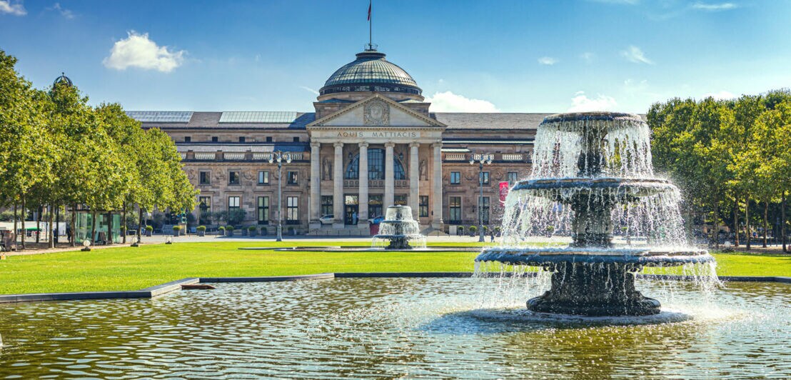 Außenansicht des historischen Kurhauses unter blauem Himmel und vor einer gepflegten Grünanlage mit Brunnen und Bäumen.