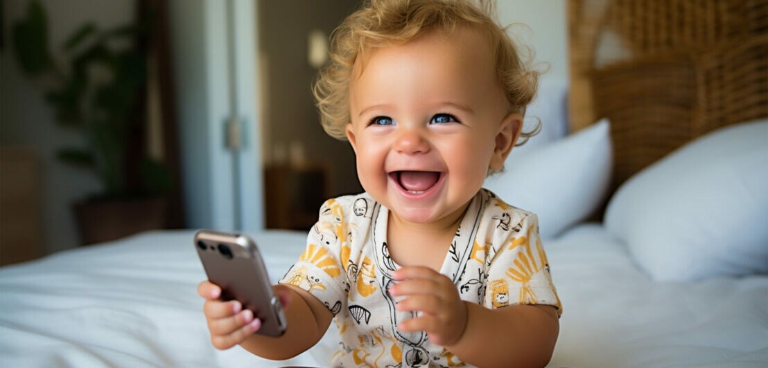 Ein Kleinkind lächelt breit in die Kamera und hält ein Smartphone in der Hand.