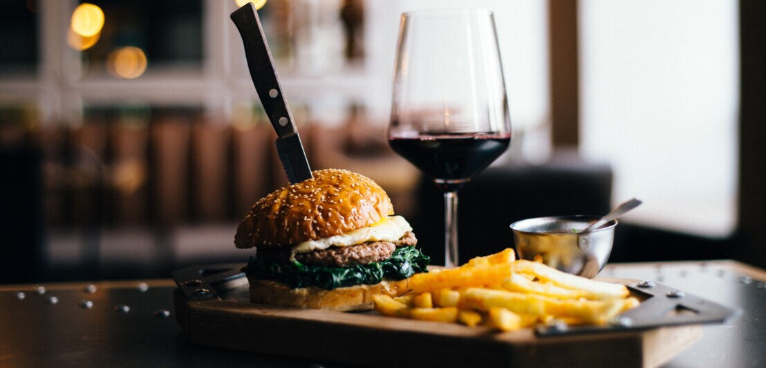 Burger mit Sesambrötchen, Spiegelei und Spinat, Messer steckt im Burger, daneben Pommes und ein Glas Rotwein auf einem Holzbrett