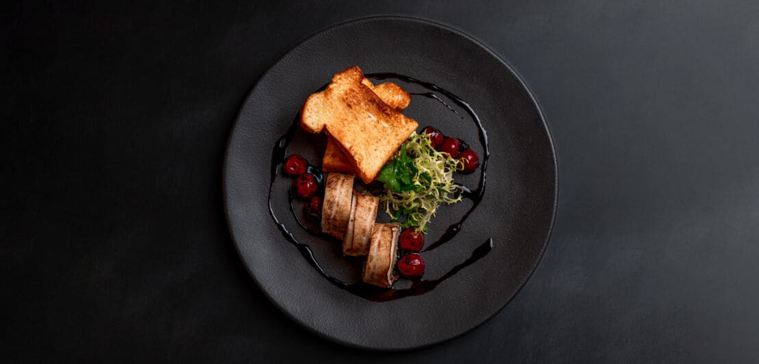 Schwarzer Teller mit zwei Scheiben geröstetem Brot, gegrilltem Fleisch, Salat und roten Beeren, angerichtet mit dunkler Sauce
