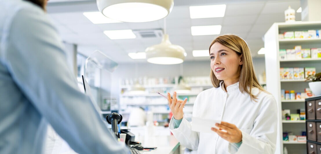 Apothekerin in weißem Kittel berät Kunden an der Apothekentheke in moderner Apotheke.