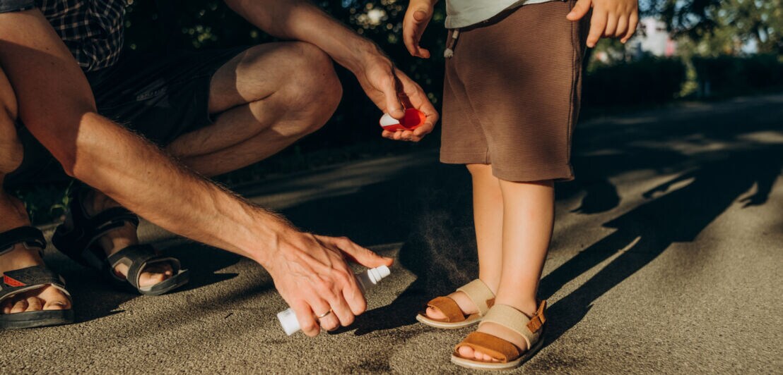 Ein Erwachsener sprüht Insektenspray auf die Füße eines Kindes.