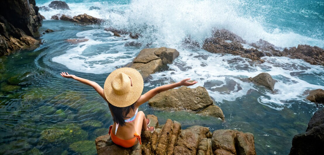 Frau mit Strohhut und orangefarbenem Bikini sitzt auf Felsen am Meer, brechende Wellen und bewölkter Himmel im Hintergrund