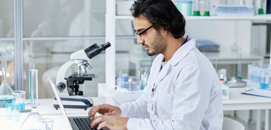 Ein Wissenschaftler sitzt im Labor an seinem Schreibtisch und arbeitet am Laptop.