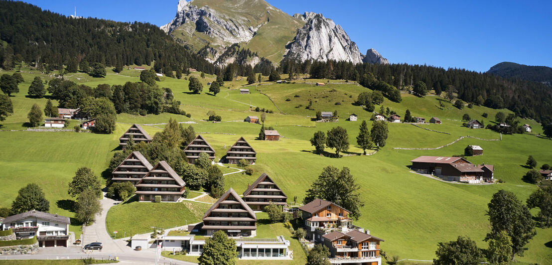 Berglandschaft mit schneebedeckten Gipfeln, grünen Wiesen und verstreuten Bauernhäusern im Vordergrund. Wolken am Himmel.