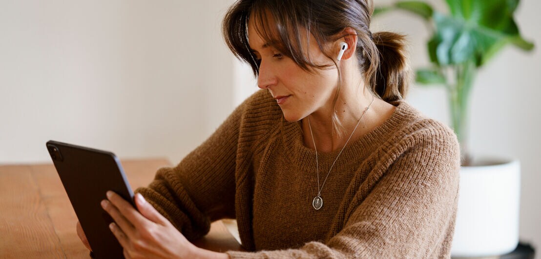 Eine Frau sitzt in an einem Tisch aus Holz und betrachtet etwas auf ihrem Tablet.