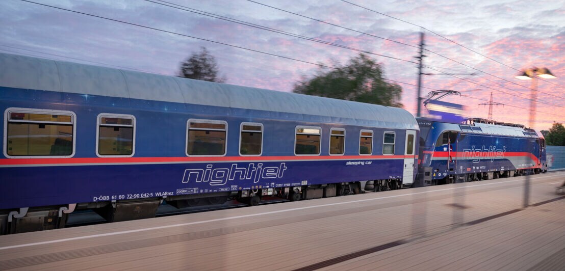 Nightjet-Schlafwagen und Lokomotive auf einem Bahnhof bei Sonnenuntergang.