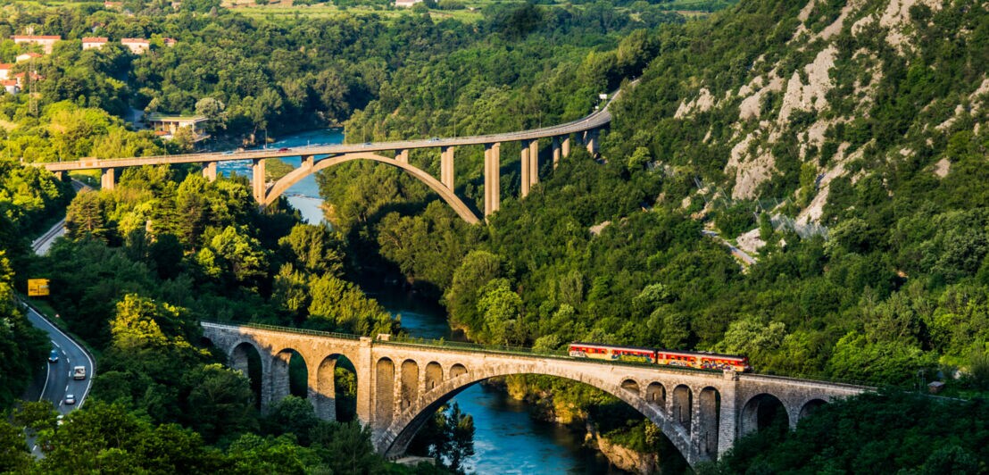 Regionalzug fährt über einen steinernen Viadukt über einen Fluss in einer grünen, bergigen Landschaft in Slowenien.