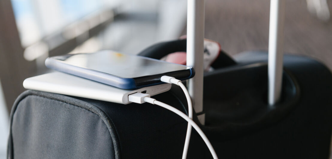 Ein Smartphone ist mit einem weißen Ladekabel mit einer Powerbank verbunden, beide liegen auf einem schwarzen Rollkoffer mit Teleskopgriff.