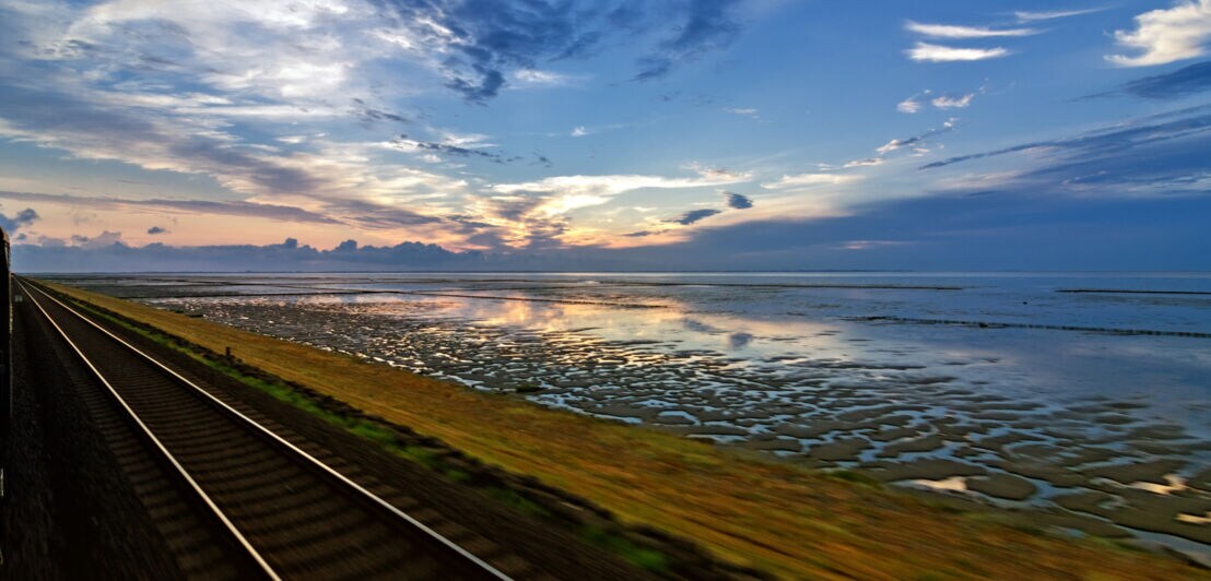 Blick auf den Hindenburgdamm, mit Schienen im Vordergrund und einem weiten Himmel mit Wolken und Sonnenuntergang im Hintergrund.