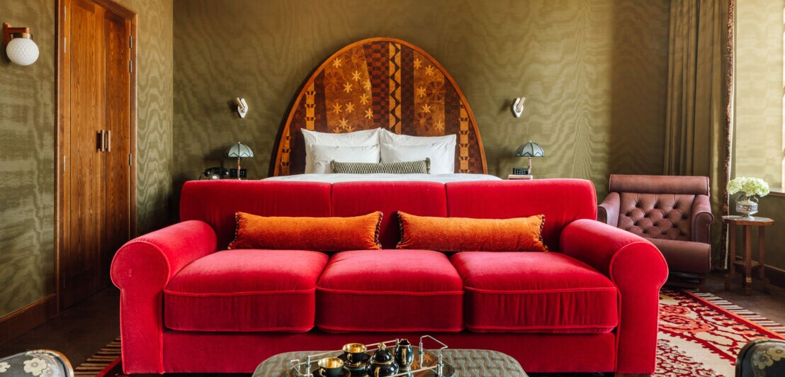 Rotes Sofa mit orangefarbenem Kissen vor einem Bett mit rundem, gemustertem Kopfteil in einem Raum mit grünen Wänden und braunen Holztüren.