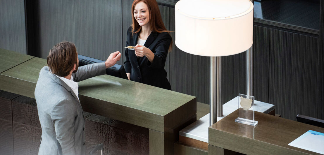 Eine Person gibt einer anderen Person über einen Tresen hinweg eine Kreditkarte an einer Hotelrezeption.