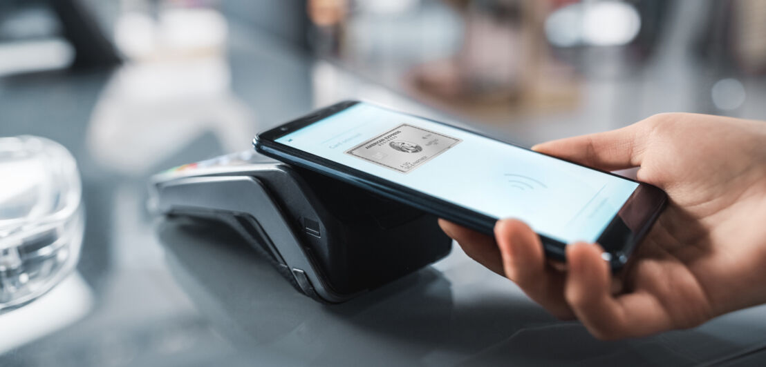 Eine Person hält ein Smartphone, dessen Bildschirm eine Kreditkarte von American Express zeigt, über ein Kartenlesegerät.