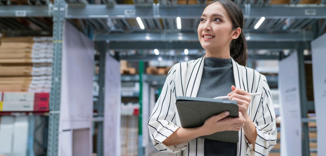 Eine Frau steht mit einem Tablet in einer Lagerhalle und hält in einer Hand einen Stift.