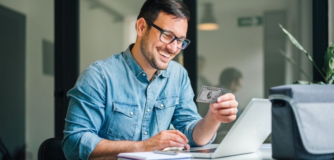 Ein lächelnder Mann im Jeanshemd bedient mit einer Hand einen Laptop auf einem Schreibtisch, in der anderen hält er eine Business-Kreditkarte von American Express.