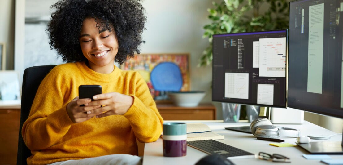 Eine Frau im gelben Pullover sitzt an einem Schreibtisch mit Monitoren und blickt lächelnd auf ein Smartphone in ihren Händen.