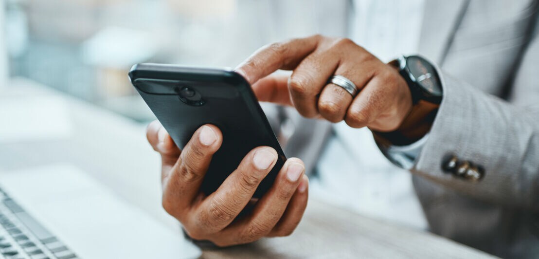 Eine Person hält mir einer Hand ein Smartphone und tippt mit dem Finger der anderen Hand auf das Display über einem Tisch mit Laptop.