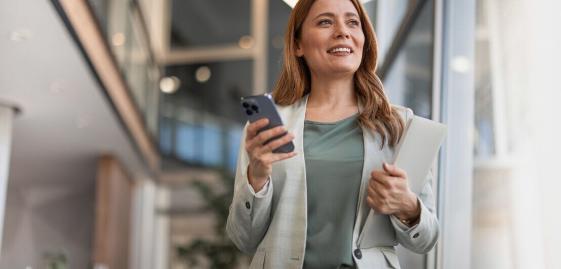 Eine lächelnde, elegant gekleidete Frau steht mit Tablet und Smartphone in den Händen in einem hellen Flur.