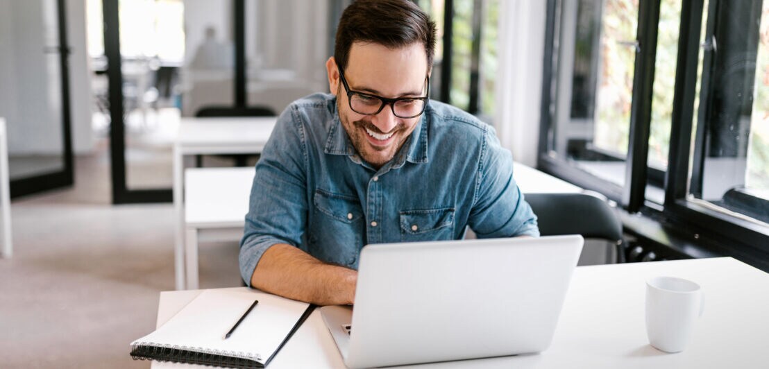 Ein Mann im Jeanshemd sitzt lächelnd vor einem weißen Laptop an einem Tisch im Büro.