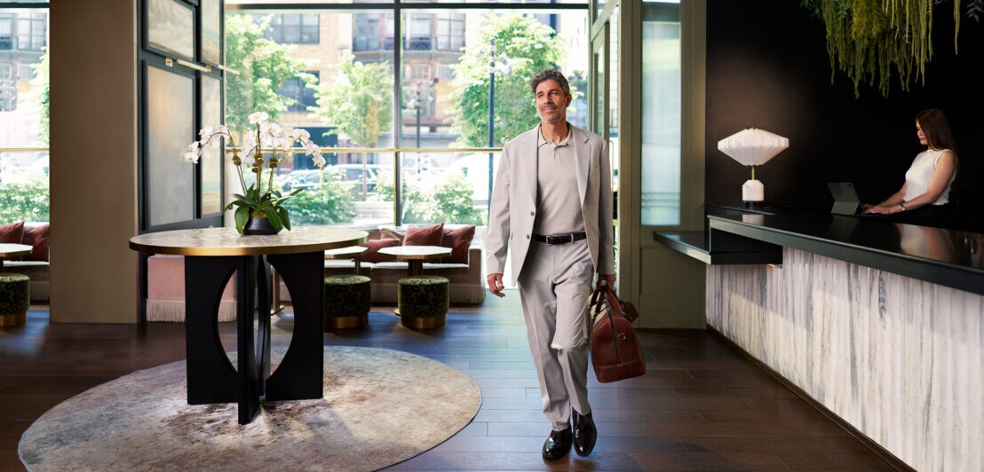 Ein stilvoll gekleideter Mann im hellen Anzug läuft lässig mit Tasche durch eine moderne Hotellobby.