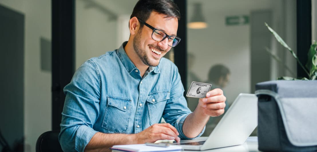 Ein lächelnder Mann im Jeanshemd sitzt vor einem Laptop und hält eine Business Platinum Card von American Express in der Hand.