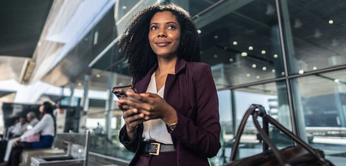 Eine Frau sitzt lächelnd mit Smartphone in den Händen vor einer Glaswand und blickt in den Raum.