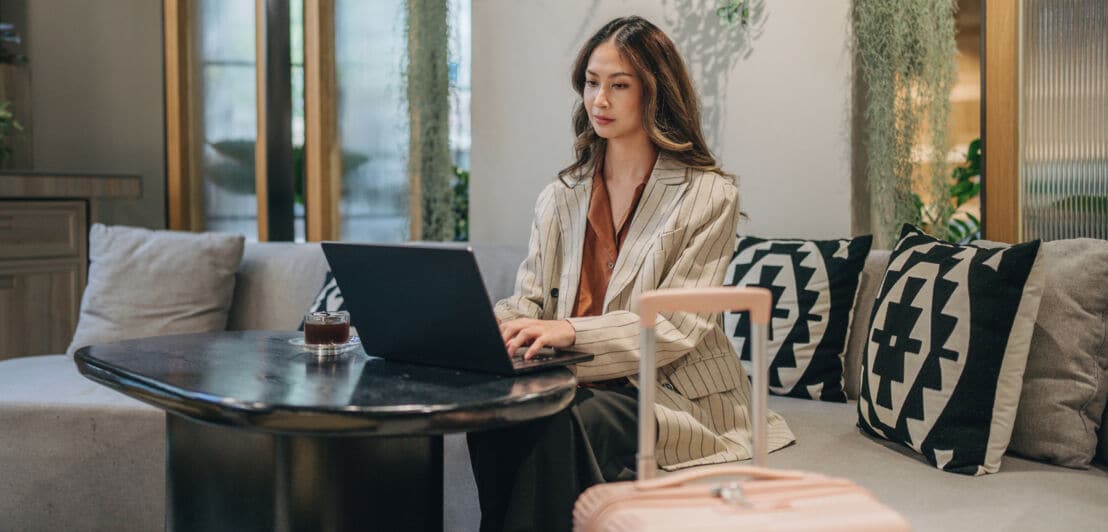 Eine junge Frau sitzt auf einem Sofa an einem Tisch und einem Laptop, neben ihr ein Hartschalenkoffer.