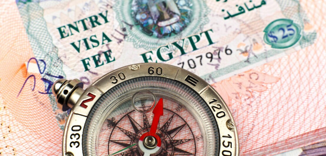 Nahaufnahme eines Kompasses mit rotem Zeiger vor einem ägyptischen Visum mit dem Text 'ENTRY VISA FEE EGYPT' und arabischer Schrift.