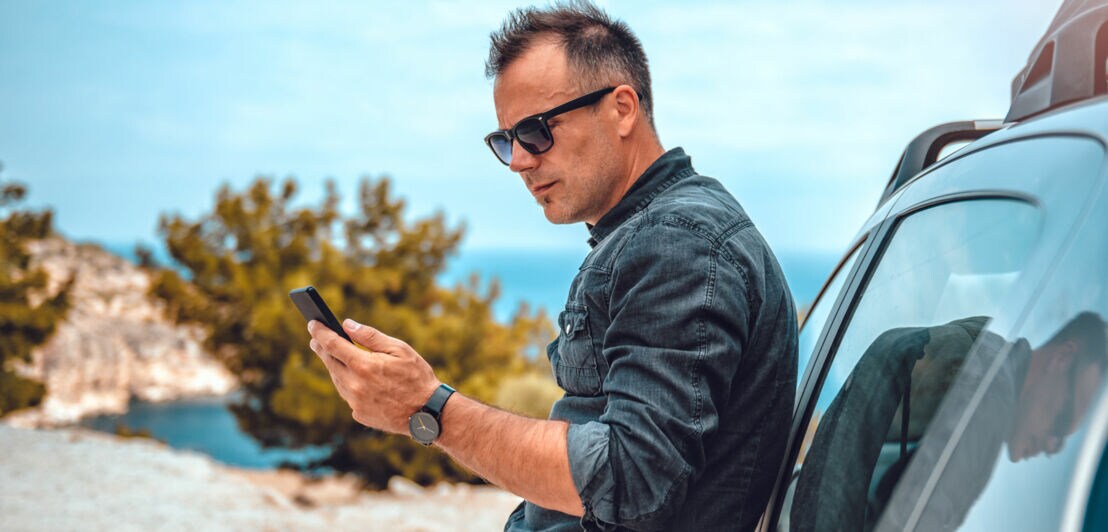 Mann lehnt an silbernem Auto und hält Smartphone in der Hand, trägt dunkles Hemd und Armbanduhr, im Hintergrund Felsen und Bäume unter blauem Himmel.