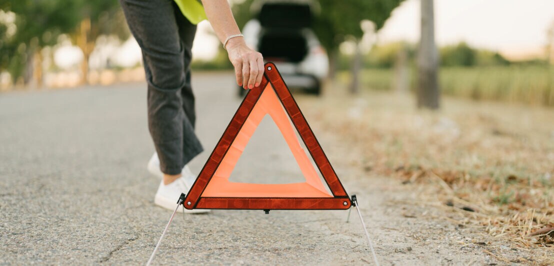 Person stellt ein rotes, dreieckiges Warndreieck mit reflektierendem Rand auf eine asphaltierte Straße.