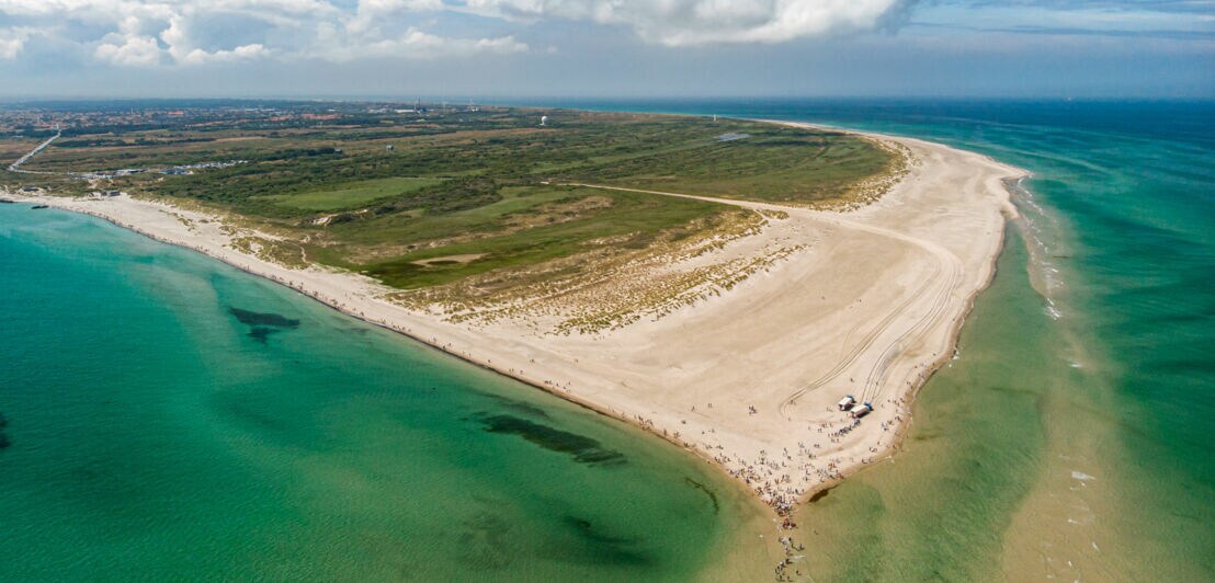 Luftaufnahme einer schmalen Landzunge mit Sandstrand und grüner Vegetation, umgeben von blauem Meer unter bewölktem Himmel.
