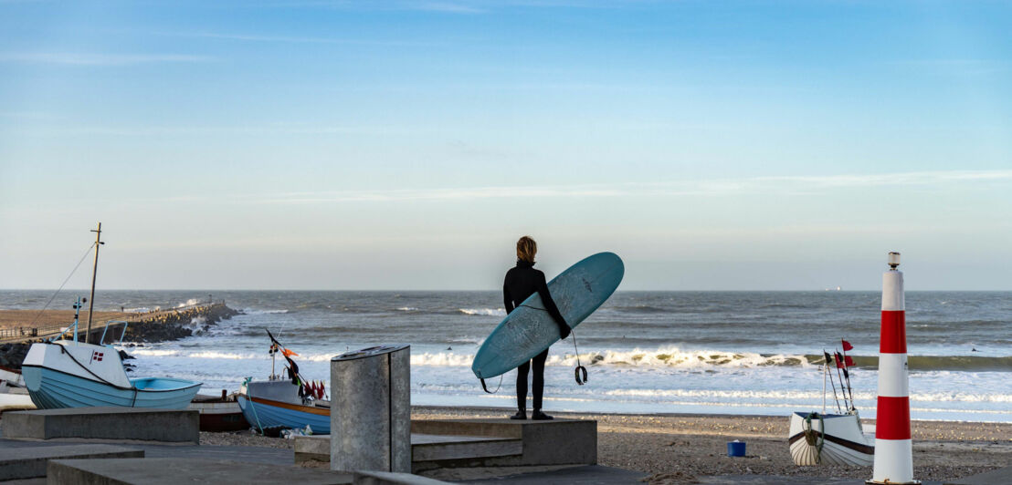 Surfer mit blauem Surfbrett steht am Strand von Dänemark und blickt auf die Nordsee, im Hintergrund Boote und ein rot-weiß gestreifter Leuchtturm.