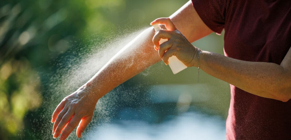 Ein Mann im T-Shirt besprüht seinen Unterarm mit Flüssigkeit aus einer Plastikflasche bei Sonnenschein.