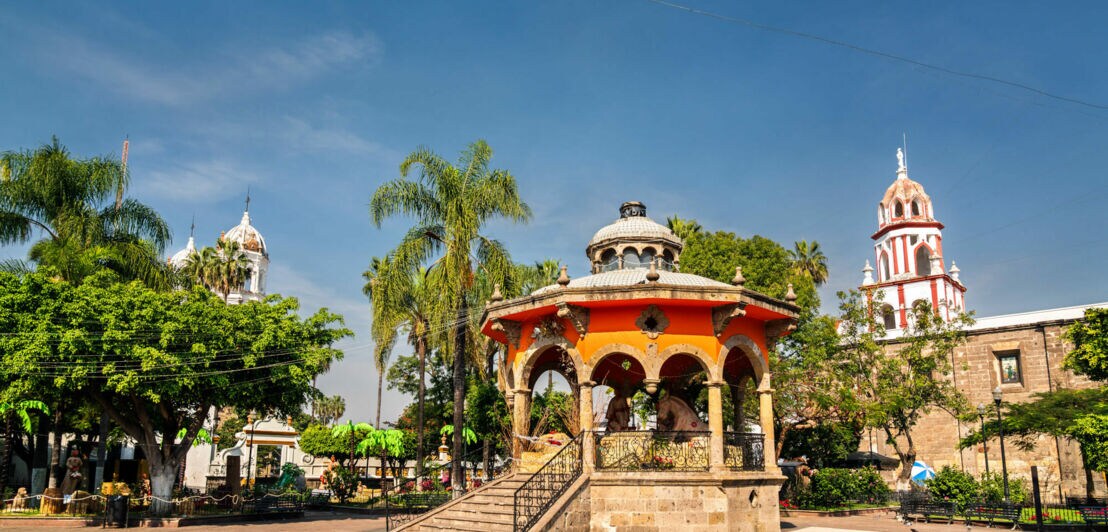 Ein Kiosk auf einem Platz mit viel Grün, im Hintergrund der Turm einer weiß-roten Kirche und blauer Himmel.
