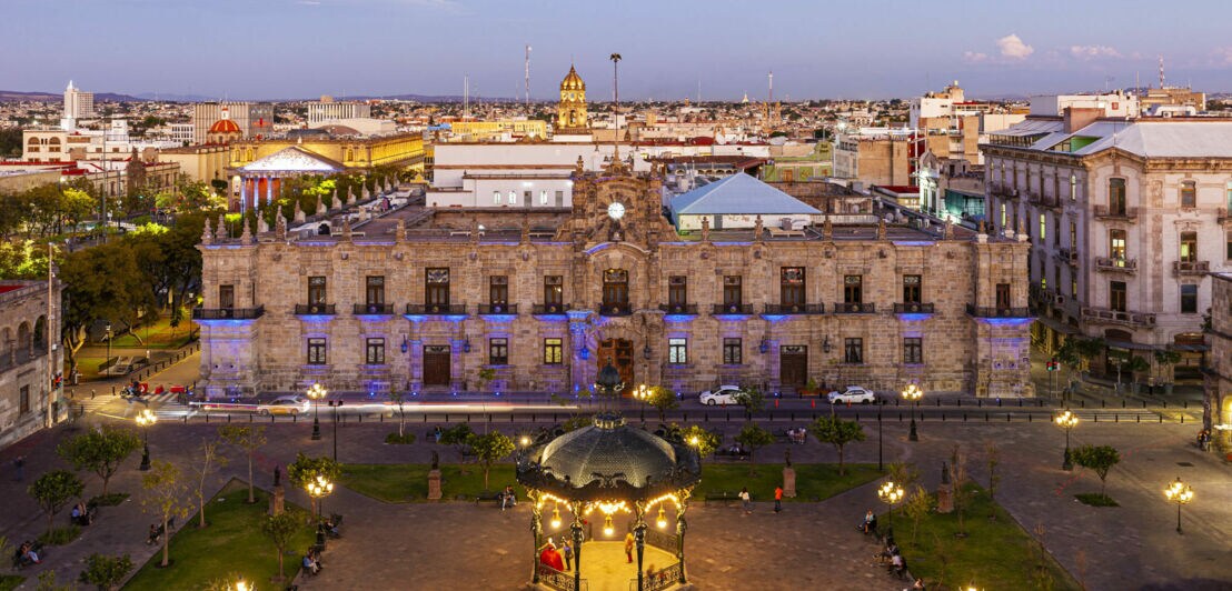 Blick von oben auf einen beleuchteten Platz vor einem prächtigen Gebäude, im Hintergrund Häuser der Stadt.