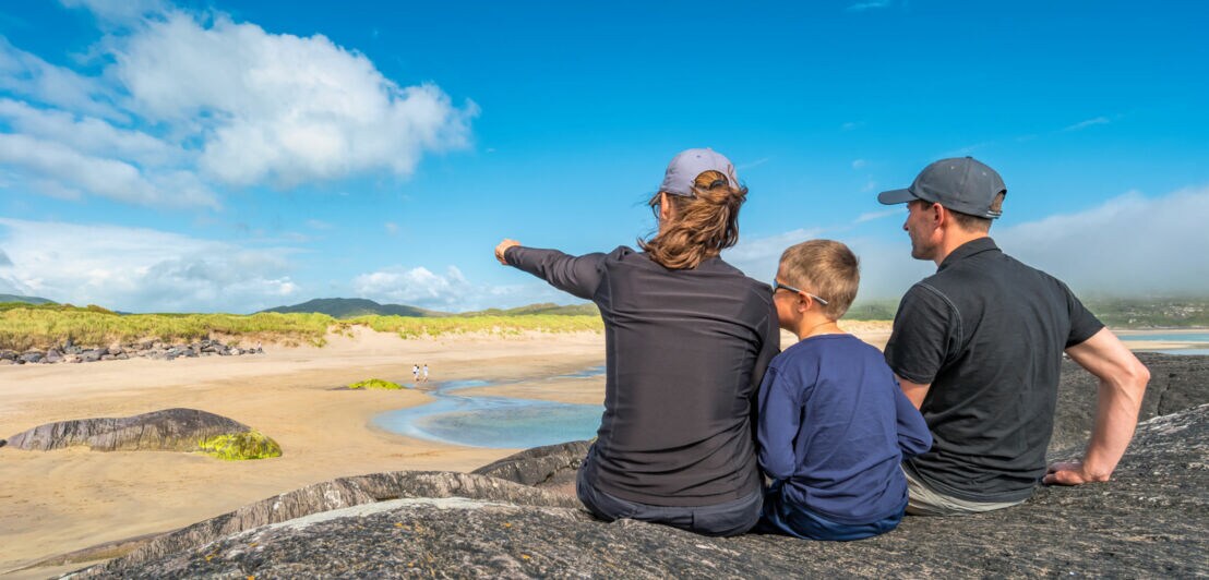 Drei Personen sitzen auf einem Felsen und blicken auf einen Sandstrand mit Dünen und blauem Himmel in Irland.