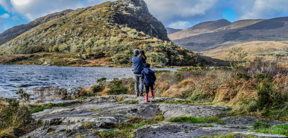 Vater mit Kindern stehen in Outdoor-Kleidung an einem Seeufer mit felsigem Untergrund, im Hintergrund bewachsene Hügel und Berge unter bewölktem Himmel.