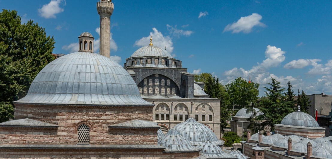 Dach eines türkischen Bads mit unterschiedlichen Kuppeln vor einer Moschee unter blauem Himmel.