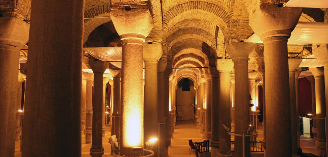 Beleuchtete, trockene Zisterne mit Sitzgelegenheiten zwischen Säulen.