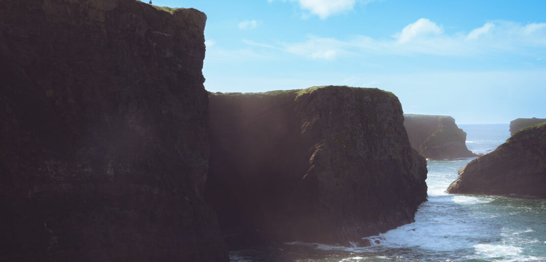 Hohe Klippen an der Küste von Kilkee mit Wellen, die gegen die Felsen schlagen, unter blauem Himmel.