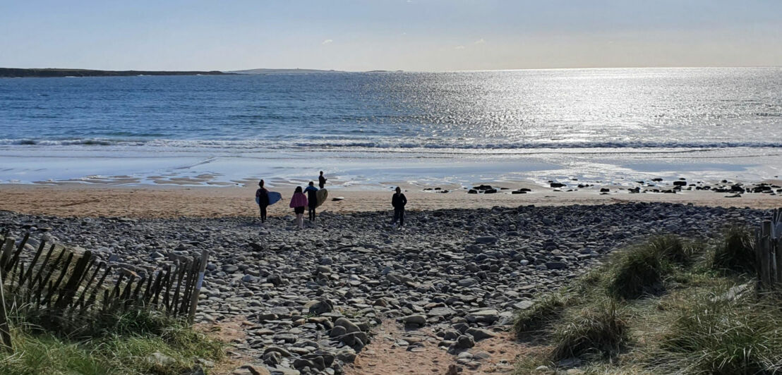 Blick auf den Strand von Spanish Point Beach mit mehreren Personen, die mit Surfbrett zum Wasser gehen, felsigem Ufer und reflektierender Meeresoberfläche bei klarem Himmel.