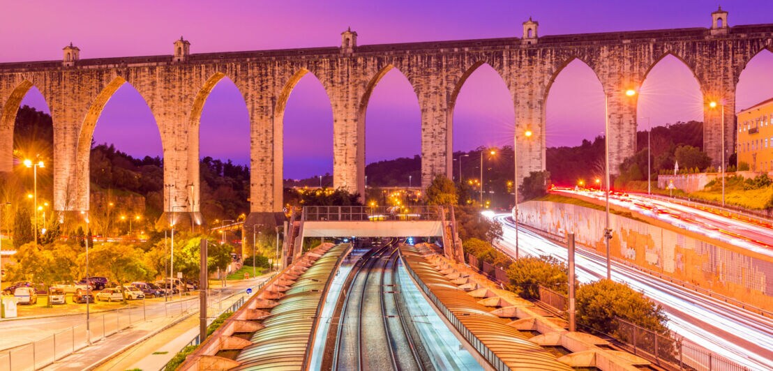Blick auf das historische Aqueduto das Águas Livres am Bahnhof Campolide am Abend mit einem surreal wirkenden Himmel im Lilatönen.