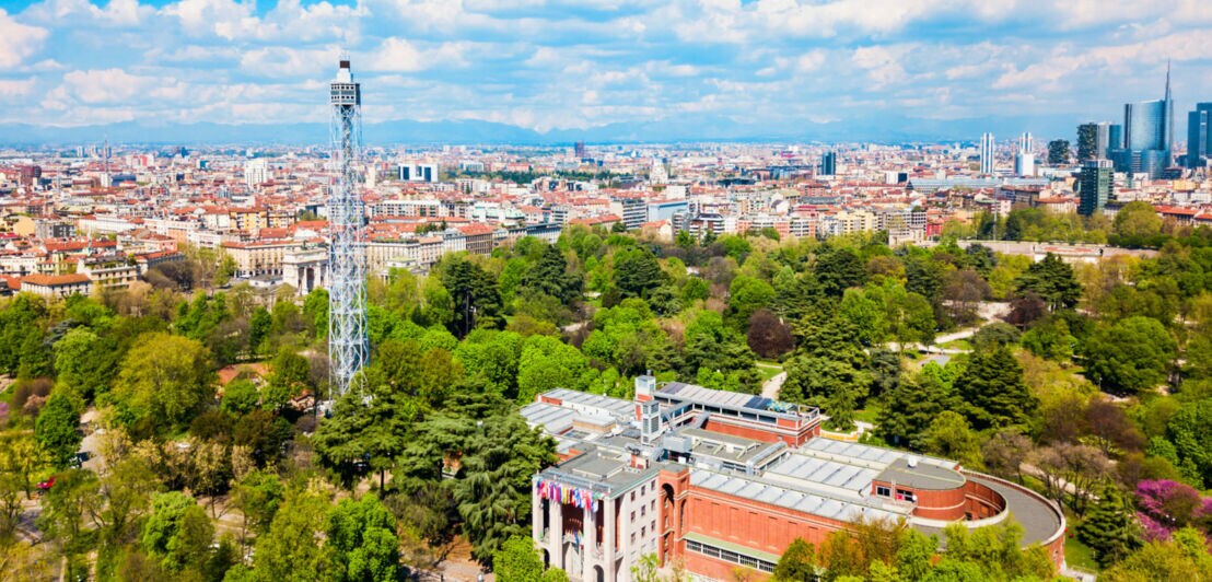 Blick von oben auf einen grünen Park mit einem großen Gebäude und einem hohen Stahlturm in der Mitte, im Hintergrund die Häuser einer Stadt.
