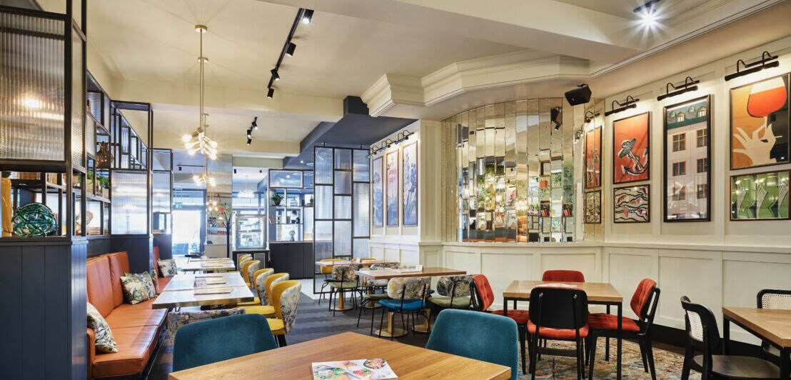 Innenraum eines Cafés mit Holztischen, gepolsterten Stühlen in Blau, Rot und Schwarz, Spiegelwand und moderner Beleuchtung.