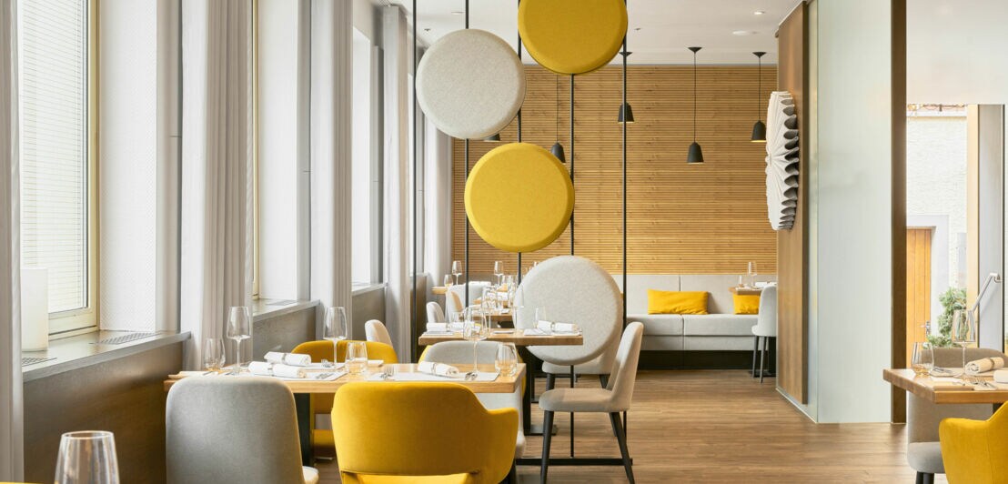 Modern eingerichteter Gastraum mit gelben und grauen Stühlen, Holzwänden und runden, schwebenden Akustikelementen in Gelb und Grau.