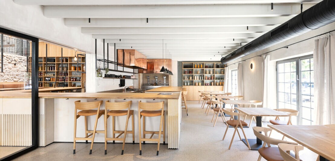 Helles Restaurant mit Holzstühlen und Tischen, offener Decke mit sichtbaren Balken, großer Fensterfront und Regalen mit Büchern im Hintergrund.