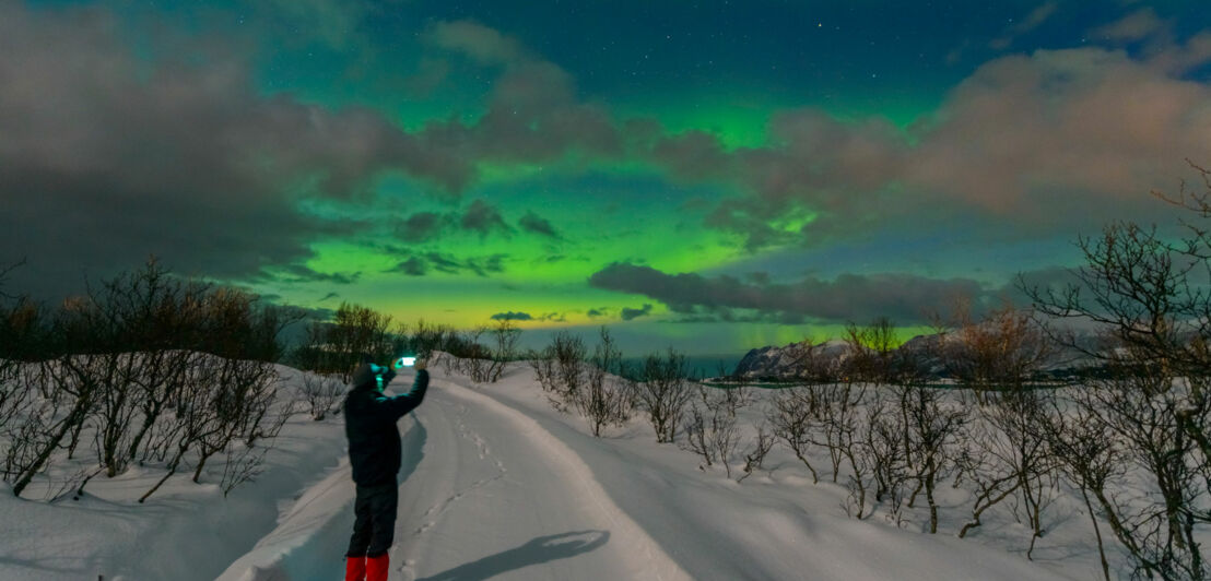 Person mit roter Hose steht auf verschneitem Weg und fotografiert grün leuchtende Nordlichter am Nachthimmel in Norwegen.