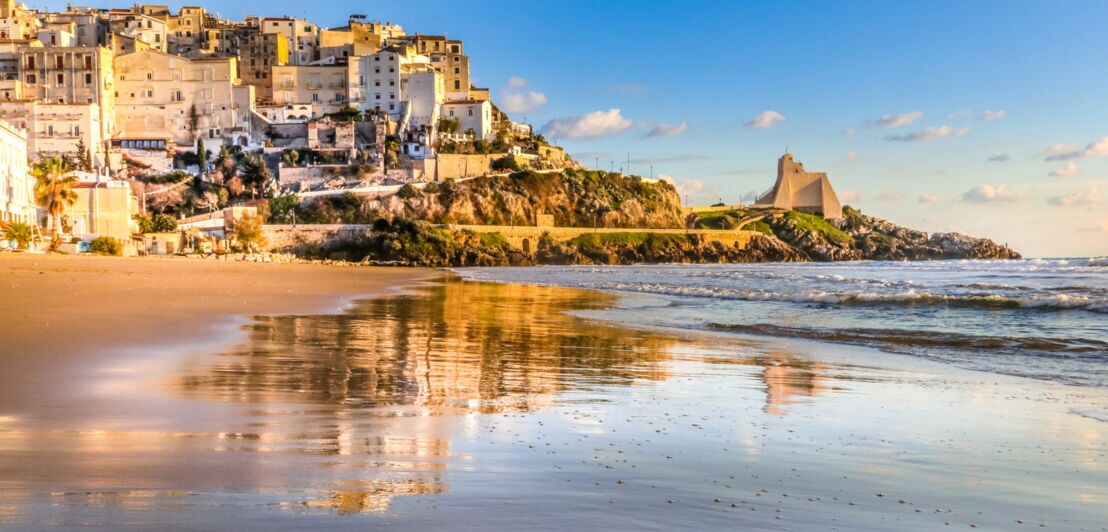 Strand mit nassem Sand, der die Häuser von Sperlonga und einen Turm auf einer Klippe im Sonnenlicht spiegelt.