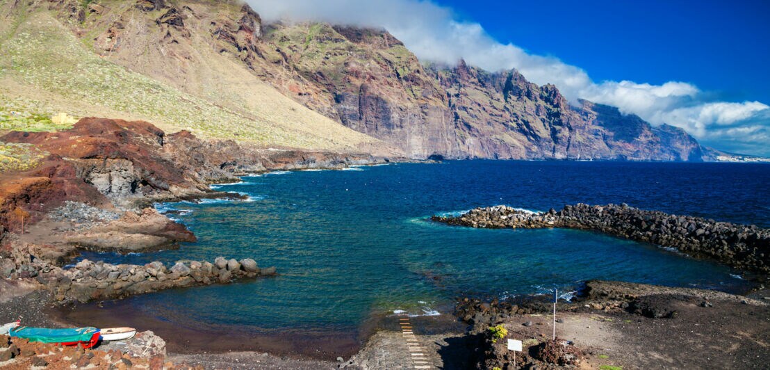 Küstenlandschaft auf Teneriffa mit steilen Felsen, blauem Meer und bewölktem Himmel, im Vordergrund ein kleiner Steg und felsige Uferbereiche.