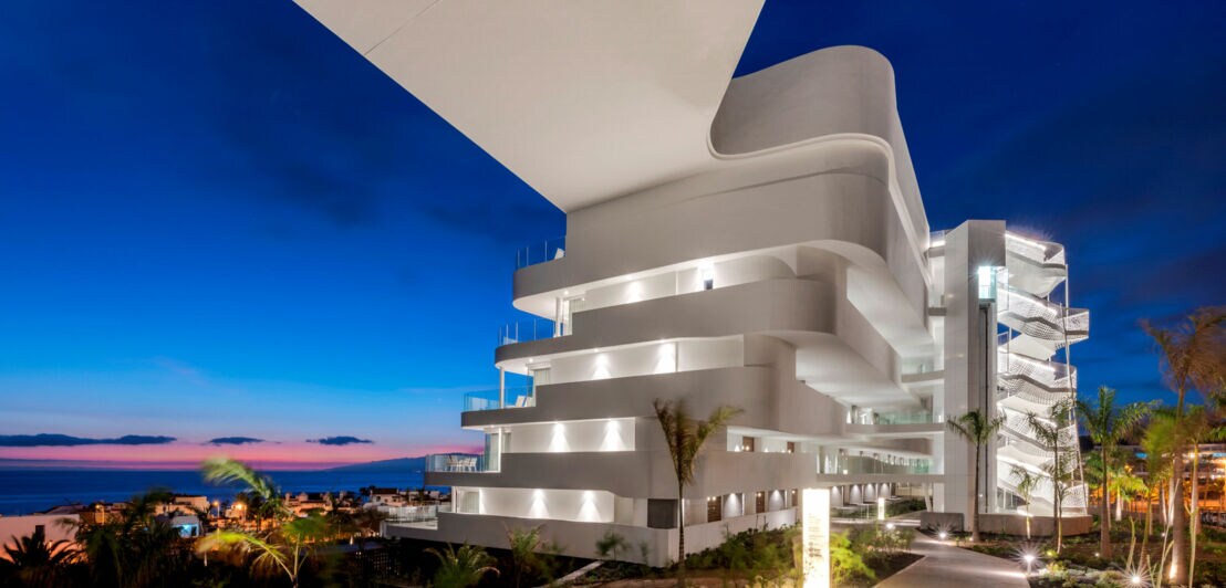 Modernes, weißes Gebäude mit geschwungenen Balkonen bei Nacht, beleuchtet, mit Blick auf das Meer und Palmen auf Teneriffa.